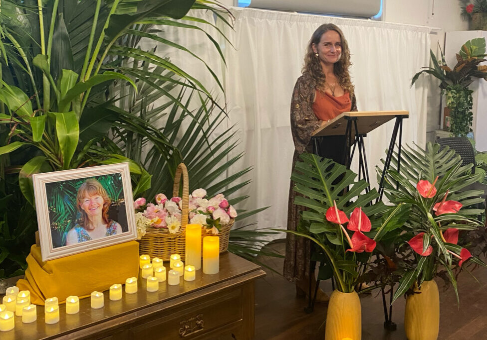 Susie Figgis in front of a memorial display with candles and framed photo, delivering a reverent eulogy in Byron Bay.